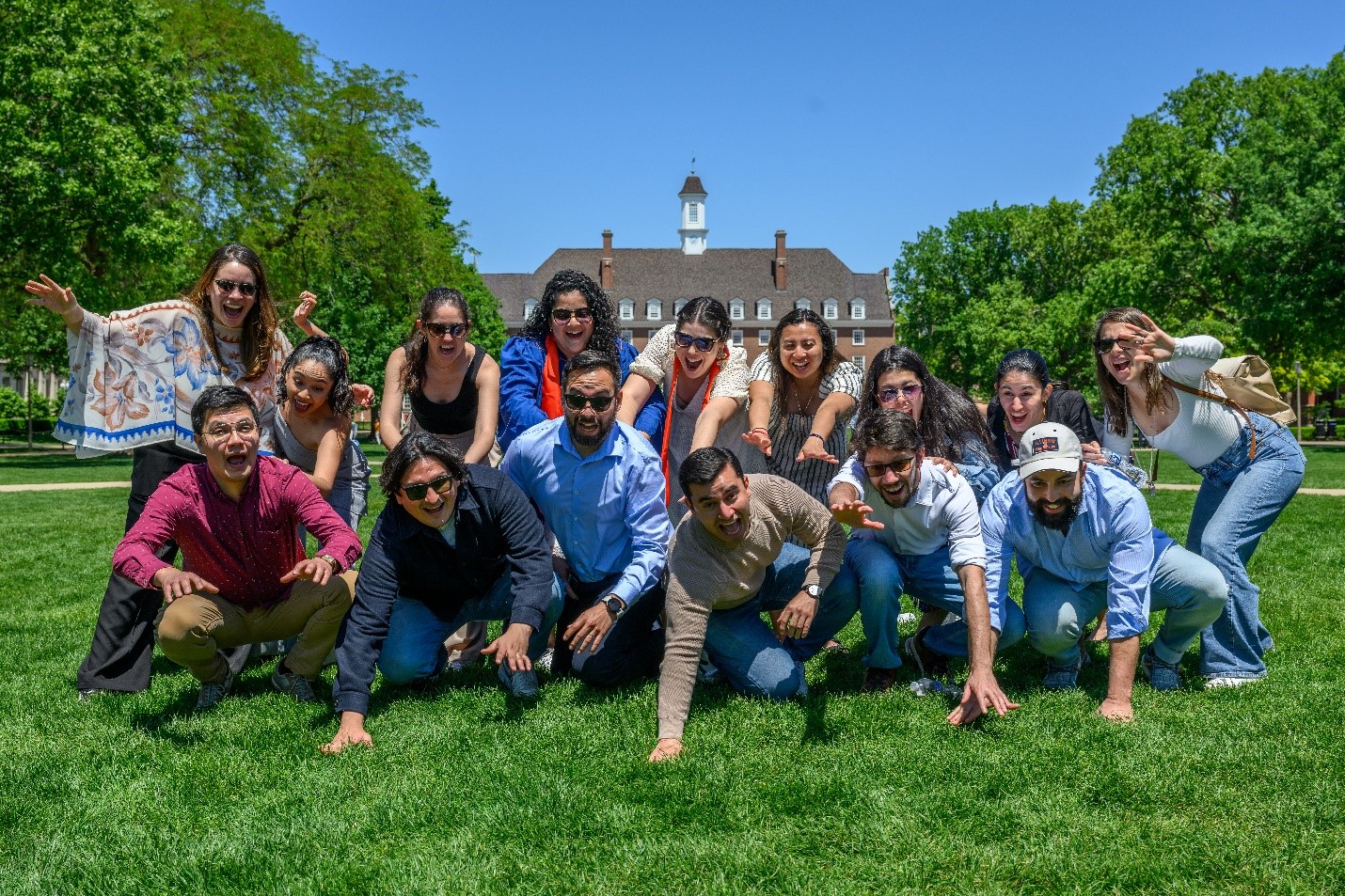 A group posing on a grassy lawn in front of a large brick building with a clock tower under a clear blue sky