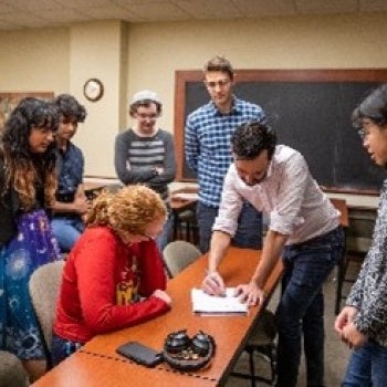 Six people in a classroom gathered around a table; one seated in a red sweater, others standing and interacting with papers. Chalkboard and desks in the background.