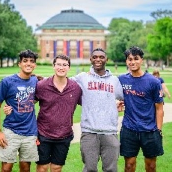 Four people standing on a grassy area with arms around each other, smiling in front of a large domed building with columns, likely on a university campus.