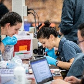 Group of people working in a laboratory setting with microscopes, laptops, and lab equipment on tables