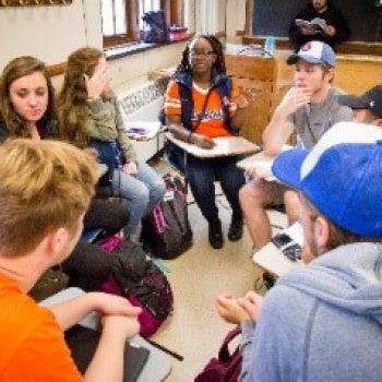 Group of people seated closely in a classroom with notebooks and backpacks visible; chalkboard and wooden desk in the background.