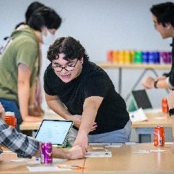 Group of people collaborating at a table in a modern workspace. One person is leaning forward and pointing at papers on the table, while others are seated and engaged in discussion. Laptops, drinks, and colorful objects are visible in the background