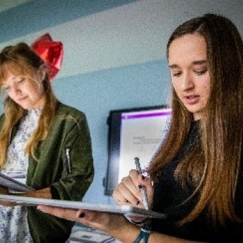 Two people standing and writing on clipboards; one wearing a light dress with a green jacket, the other in a dark top. A whiteboard or screen with unclear text is in the background