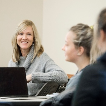 Two students sitting at desks in a classroom setting. An instructor sits at a separate desk and smiles 