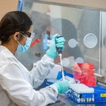 Person in a laboratory wearing protective gear—lab coat, gloves, goggles, and mask—using a pipette to transfer liquid into test tubes in a blue rack, with lab equipment in the background