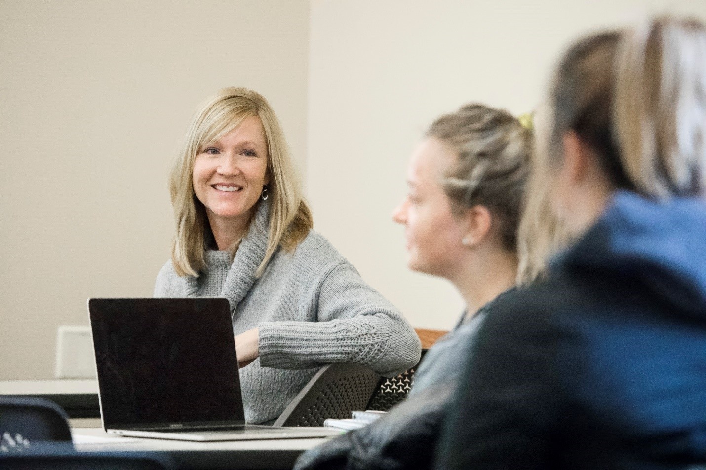 Two students sitting at desks in a classroom setting. An instructor sits at a separate desk and smiles 