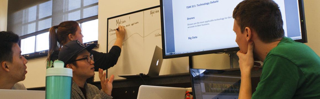 A group of students work together in a classroom setting, using a whiteboard and large display monitor